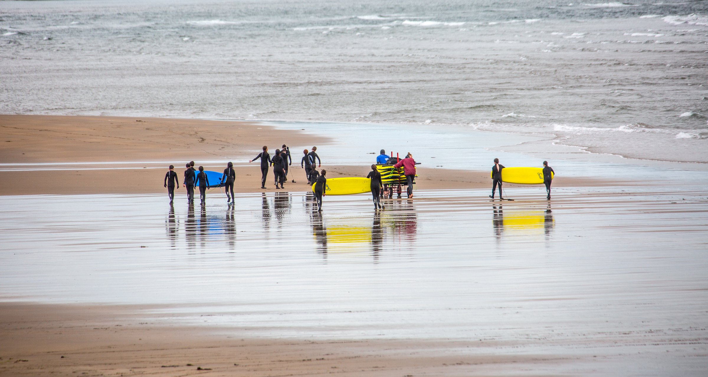 The whole photo: the two surfers are part of a surf class of about fifteen young people in wetsuits, walking to the sea with their boards while two adults push a trolley of boards