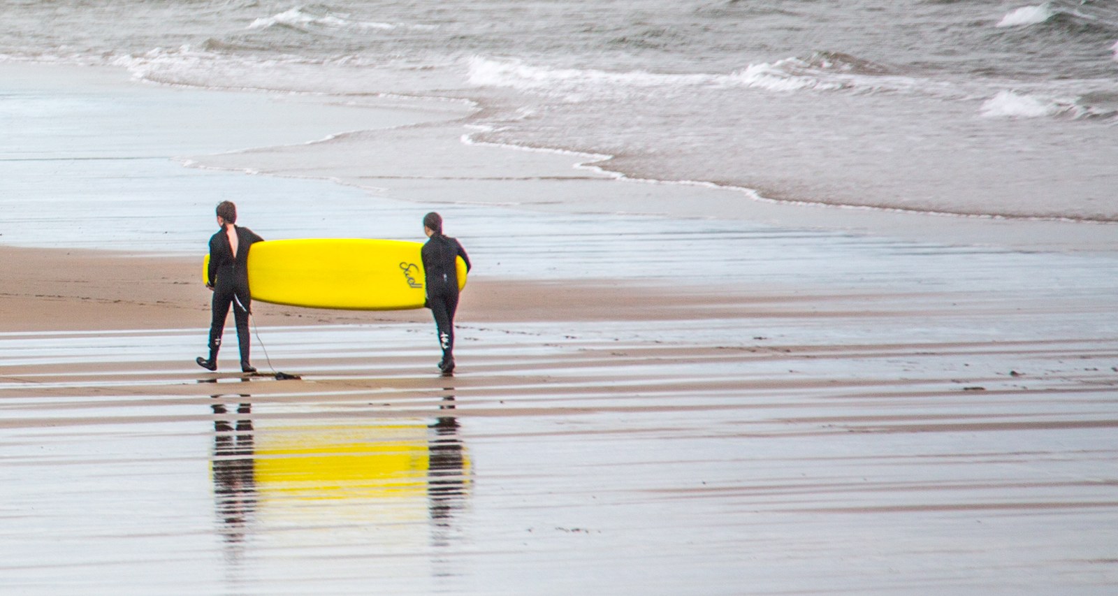 Two surfers in wetsuits carry one yellow surfboard towards the sea, alone on a wide, empty beach